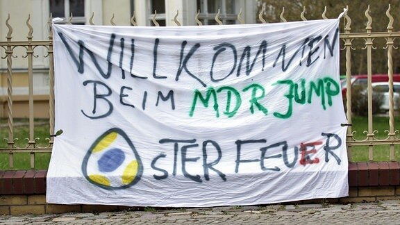 An einem Zaun hängt ein selbstgestaltetes Plakat mit der Aufschrift "Willkommen beim MDR JUMP Osterfeuer"