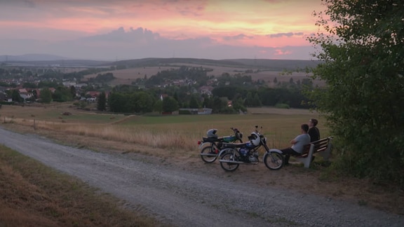 2 Simsons am Rand eines Feldes bei Sonnenuntergang. 2 Menschen sitzen daneben auf einer Bank