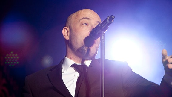 Ein Mann mit Glatze im Anzug singt in eine Mikro.