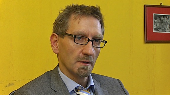Ein Mann sitzt vor einer gelbgestrichenen Wand. Er trägt einen grauen Anzug, eine schwarze Brille und hat kurzes, strubbeliges Haar sowie einen Oberlippen- und einen Kinnbart.