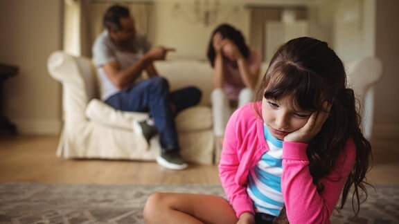 Kind schaut traurig während Eltern sich im Hintergrund streiten.