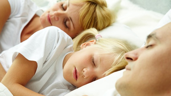 Ein Elternpaar schläft, in der mitte liegt ein blondes Kleinkind und schläft ebenfalls.