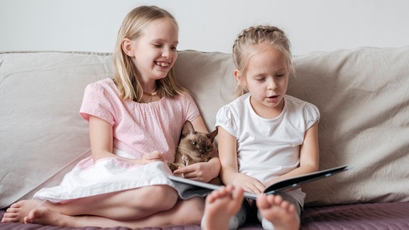 Two little girls sitting barefoot on the couch with Burmese cat watching a book model released Symbolfoto property released PUBLICATIONxINxGERxSUIxAUTxHUNxONLY EYAF00914