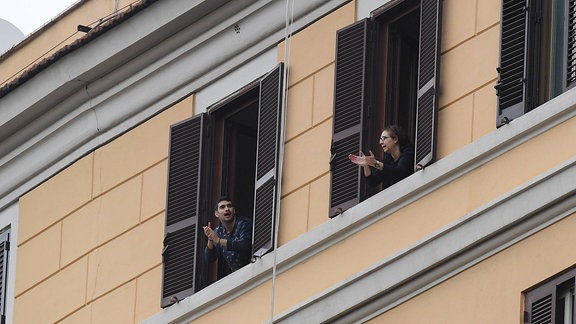 Italien: Zwei Bewohner applaudieren an den Fenstern ihrer Wohnungen in der Innenstadt von Mailand für vorbeifahrende Einsatzfahrzeuge, Ärzte und Sanitaeter