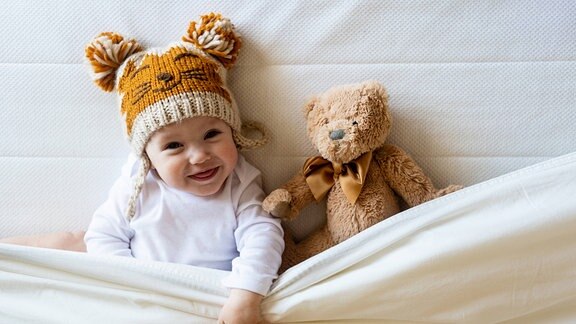 Kleiner Junge mit Mütze und Teddy im Bett (Symbolbild)