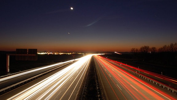 Eine Autobahn bei Nacht, die Lichter zu roten bzw. weißen Streifen verschwommen. Am Horizont geht die Sonne unter, ein kleiner Streifen Himmel schimmert noch rötlich.
