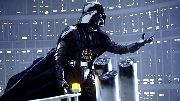 Darth Vader aus Star Wars steht auf der Brücke seines Raumschiffes und streckt den Arm empor.