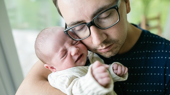 Vater mit Brille hat drückt seine Wange an schreiendes Baby, das er im Arm hält.