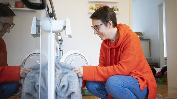 Frau in einem orangefarbenen Pullover und blauer Jeans hockt vor einer Waschmaschine und lächelt. Sie hat eine Brille auf und steckt ein hellgraues Kleidungsstück in die Maschine