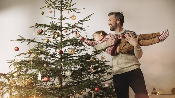 Ein Vater hebt seinen Sohn an den Weihnachtsbaum, damit dieser die Dekoration anbringen kann. Sie lachen beide.