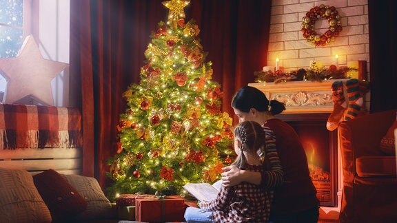 Mutter liest ihrem Kind vor Weihnachtbaum etwas vor. Merry Christmas and Happy Holidays! Pretty young mom reading a book to her cute daughter near Christmas tree indoors. 