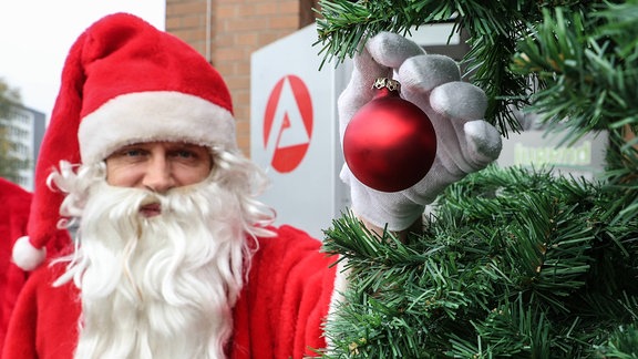 Weihnachtsmann steht an weihnachtsbaum vor Arbeitsagentur und hängt eine Weihnachtskugel auf
